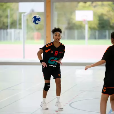 Young handball players in black and orange jerseys on an indoor court, with a ball in mid-air during a match.