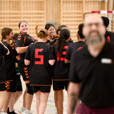 Handball players in black and orange jerseys huddle together on the court while a bearded man in black looks on.
