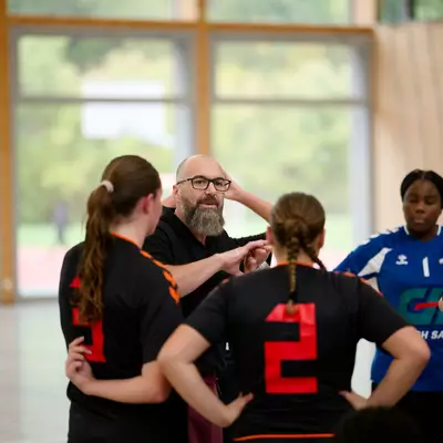 Coach gesturing while talking to players in black and orange jerseys, with a goalkeeper and teammate drinking water nearby.