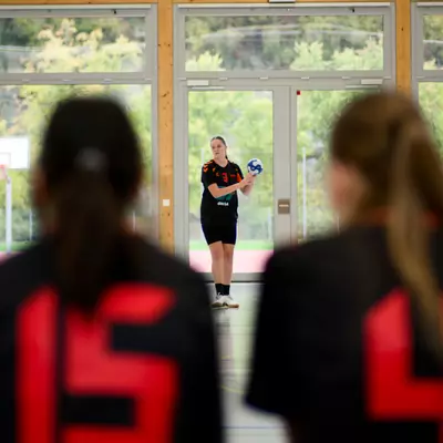 Coach holding a ball faces two players wearing numbered jerseys (15 and 4) in an indoor handball court.