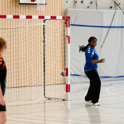 Two young female handball players practice in a gym, one in a blue jersey near the goal, the other in orange and black.