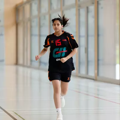 Young female handball player in black and orange jersey number 15 running across an indoor gym court.