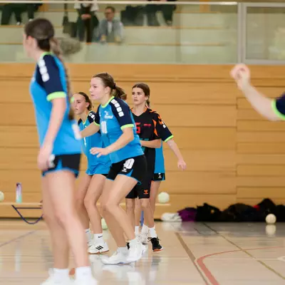 Young female handball players in light blue and dark jerseys compete on an indoor court during an M16 match.