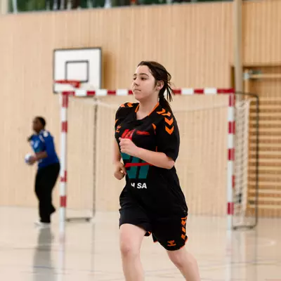 Young female handball player in black and orange jersey running on court, with a goal and referee visible behind her.