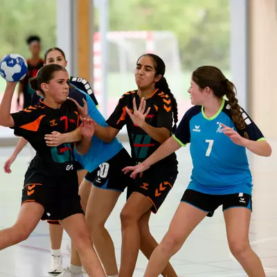 Female handball players in black and light blue jerseys compete for the ball during an indoor match.