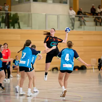 Handball player in mid-air shooting the ball while opposing defenders in blue jerseys attempt to block on an indoor court.