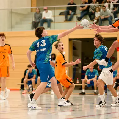 Handball players in orange and blue jerseys compete for the ball during a youth match, with player 13 attempting a shot.