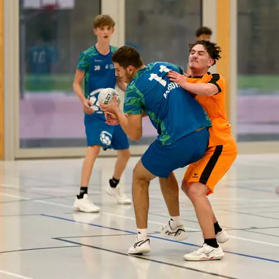 A handball player in blue holding the ball while being defended by an opponent in orange on an indoor court.