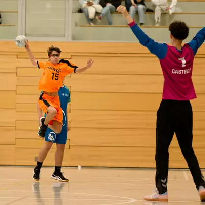 Handball player in orange jersey number 15 jumping to shoot while opponent in pink defends with arms raised.