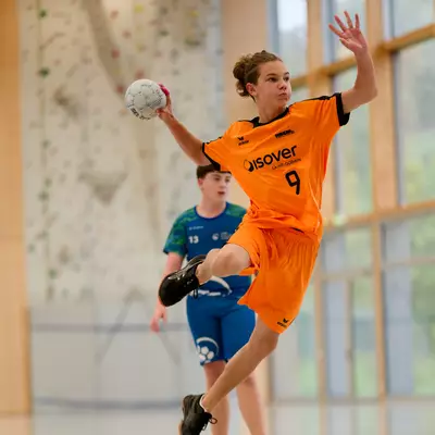 Handball player in orange jersey number 9 leaping mid-air to throw, with an opposing player in blue watching behind.
