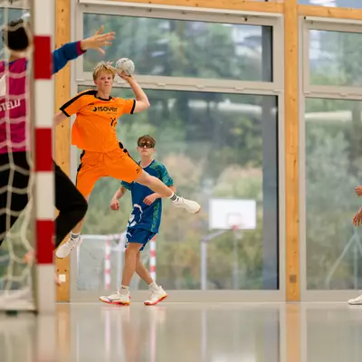 A handball player in an orange jersey jumps to shoot while a goalkeeper and teammates look on in an indoor court.