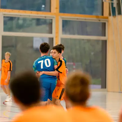 Two young handball players, one in blue (number 70) and one in orange, embrace on the court during a match.
