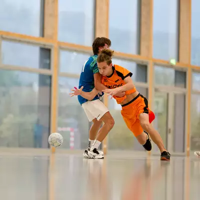 Two M17 players in blue and orange jerseys challenge for the ball during an indoor handball match.
