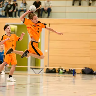 Handball player in orange jersey leaping mid-air to throw the ball during a match, teammate and goalkeeper nearby.