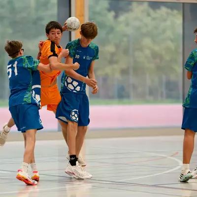Young handball players in blue and orange jerseys compete on an indoor court, with one player jumping to shoot.