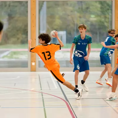 Young handball player in orange jersey number 13 leaping to shoot while defenders in blue close in during an indoor match.