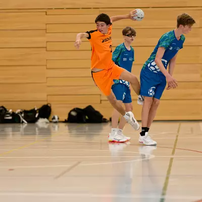 Handball player in orange jersey jumping to throw the ball, guarded by two defenders in blue on an indoor court.