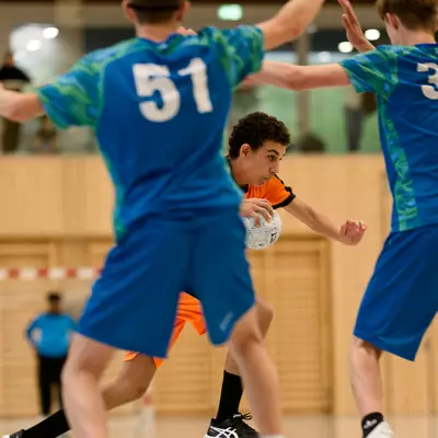 Handball player in orange jersey driving with the ball between two defenders in blue jerseys with raised arms.