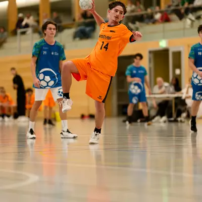 Handball player in orange jersey number 14 jumping to throw the ball during a youth M17 match on an indoor court.