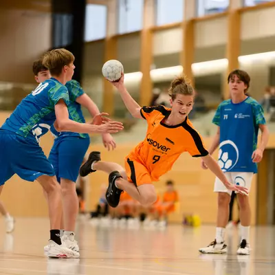 Handball player in an orange jersey jumps to shoot while defenders in blue attempt to block during an indoor match.
