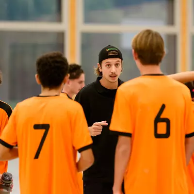 Youth handball players in orange jerseys gather around their coach during a timeout, listening to instructions.