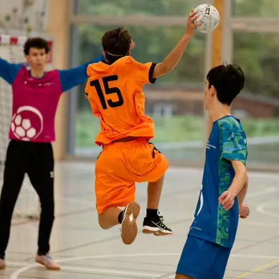 Handball player in orange jersey number 15 leaping to shoot, with a goalkeeper and defender nearby on the court.