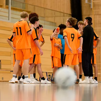 Young handball players in orange jerseys huddle on the court with their coach, a ball blurred in the foreground.