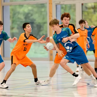 Young male handball players in orange and blue jerseys compete for the ball during an indoor match.
