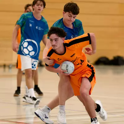 Young handball players in action on an indoor court, with a player in orange holding the ball while defended by players in blue.