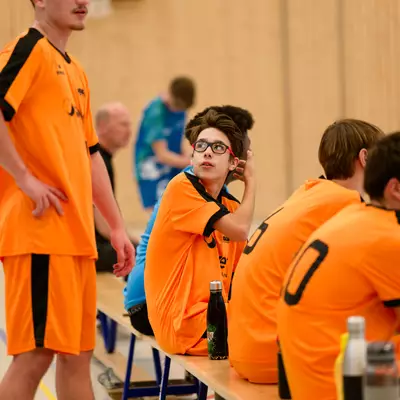 Young male handball players in orange jerseys sit on a bench, one resting his head on his hand, during a break in play.