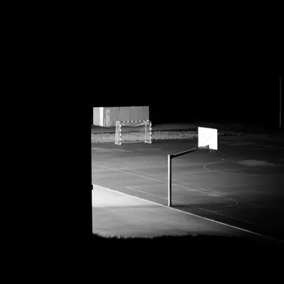 Night-time black-and-white shot of an empty outdoor basketball court, illuminated by floodlights with a goal frame and hoops visible.
