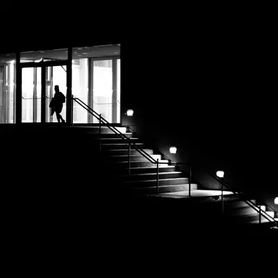 Silhouette of a person walking through a glass doorway at the top of an outdoor staircase, lit by small lamps at night.