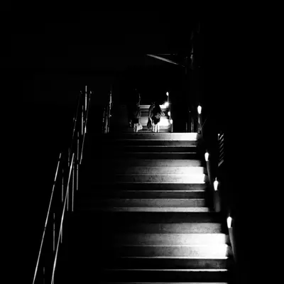 Black-and-white night photo of a dimly lit staircase with handrails, a silhouetted figure visible at the top near a faint light.