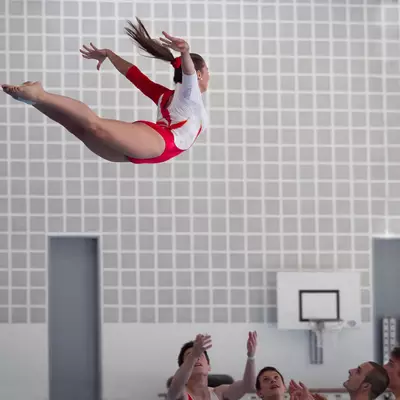 A gymnast in red and white attire performs a spectacular aerial figure, her body suspended horizontally in mid-air. Against a grey gridded wall background, her dynamic movement creates striking contrast. Below, several spotters raise their arms, ready to catch her. The vivid red of her outfit against the monochromatic environment highlights the athletic performance.
