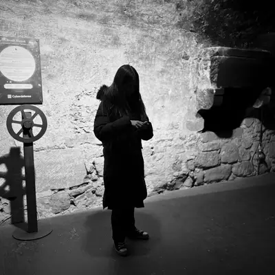 In an underground stone-walled environment, a dark figure stands near a circular sign mounted on a pole. Dramatic lighting creates strong contrast between shadows and light, isolating the silhouette against the textured wall. The black and white atmosphere enhances the scene's mysterious nature.
