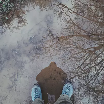 A photo taken vertically over a puddle. You can see my feet in the puddle and my reflection and the one of branches of a tree.