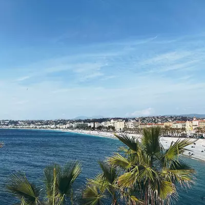 Panoramic view of the Promenade des Anglais in Nice. Palm trees in the foreground frame the view of the famous crescent-shaped bay. The pebble beach stretches along the coastline, bordered by historic buildings with characteristic ochre facades. The deep blue Mediterranean Sea contrasts with the azure sky. A few people can be seen walking on the beach.