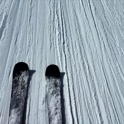 A close-up view of skis on groomed snow, photographed from above. The tips of two black skis are visible against pristine white snow that shows parallel grooming lines. The texture of the packed snow is clearly visible with its characteristic linear patterns created by the grooming machine. The image has a strong geometric quality with the parallel lines of both the ski tracks and grooming marks.