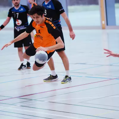 An indoor handball match in progress. A young player in an orange and black jersey (likely number 2) is handling the ball while being defended by players in blue and black uniforms (one wearing number 17). The scene takes place in an indoor sports hall with typical handball court markings visible on the floor. The image captures the dynamic movement of handball, with the attacking player in mid-motion while controlling the ball.