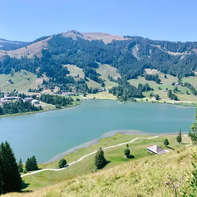 vu en surplomb du lac noir. Au centre de la photo se trouve le lac noir. Sur la rive du bas un chemin longe le lac. Sur la rive au-dessus, on peut voir le camping ainsi que le camps de vacances du village Schwarzsee Bad.