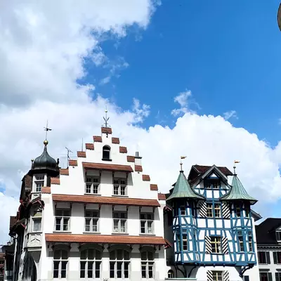 Two house fronts in the old town of St. Gallen. The one on the left has brown eaves above the windows. Its façade is white. The house on the right has two towers and blue woodwork.