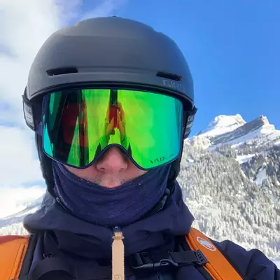 A selfie of Cedric wearing a helmet and a ski mask. The phone is reflected in the green visor of the mask.